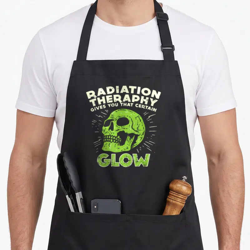 Fighter radiation aprons - Distressed green skull with white radiating lines and sarcastic text about radiation therapy — shown on a black cotton canvas apron against a white background