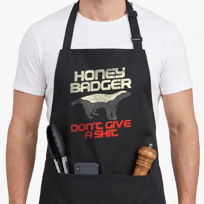 Honey give badger apron - Distressed honey badger graphic with beige and red text on grey background — shown on a black cotton canvas apron against a white background