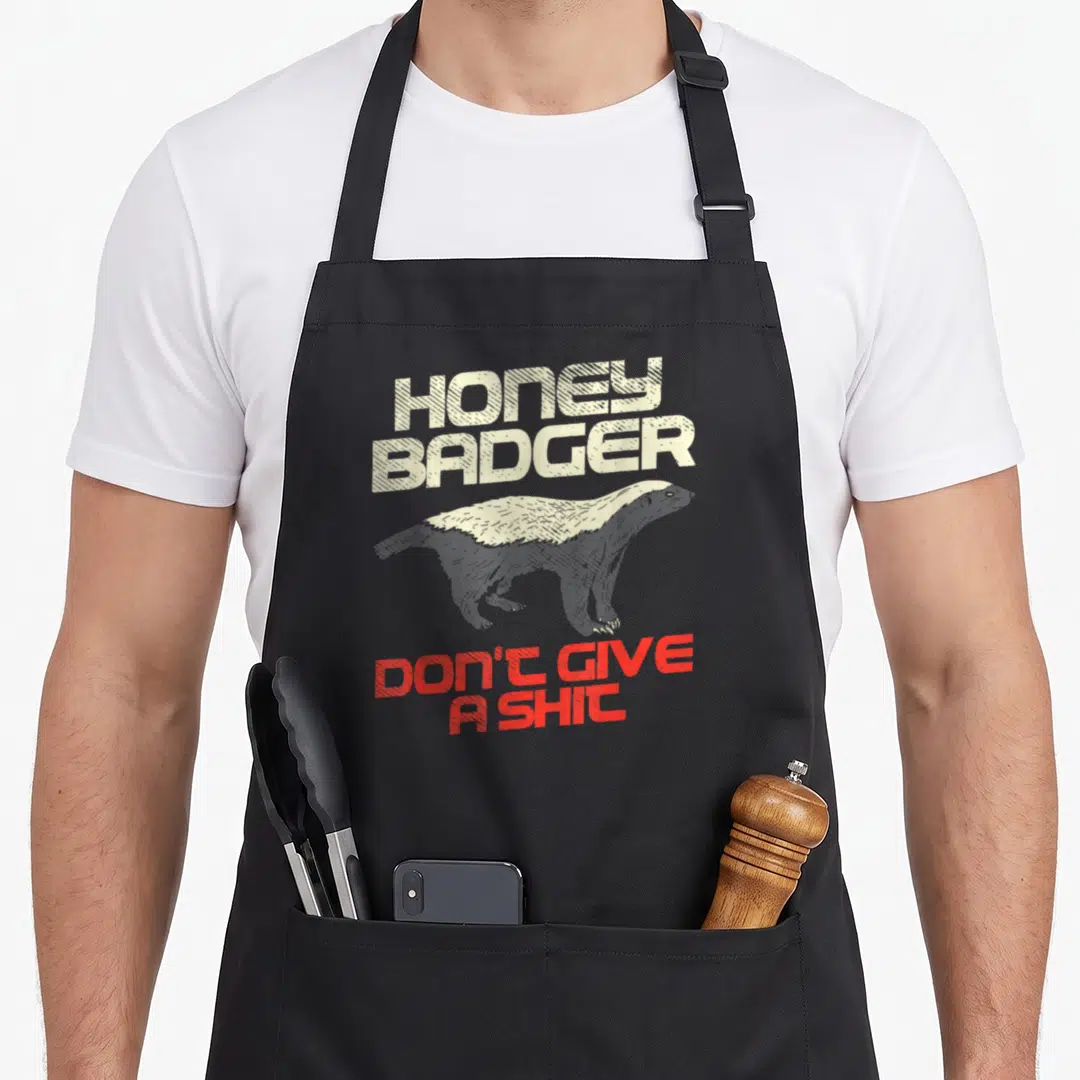 Honey give badger apron - Distressed honey badger graphic with beige and red text on grey background — shown on a black cotton canvas apron against a white background