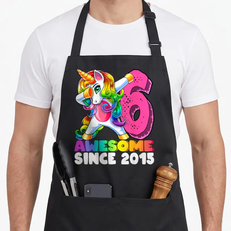 Dabbing unicorn birthday apron - Cartoon unicorn dabbing next to pink number 6 and rainbow AWESOME SINCE 2015 text — shown on a black cotton canvas apron against a white background