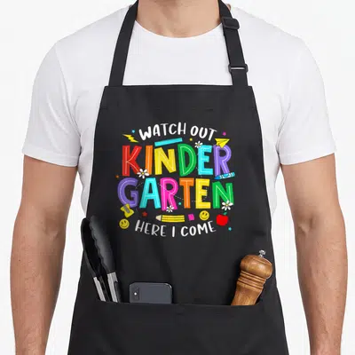 Kindergarten watch out apron - Colorful KINDER GARTEN text surrounded by pencils, apple, sharpener, and flowers — shown on a black cotton canvas apron against a white background