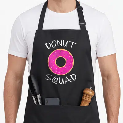 Funny squad donut apron - Pink cartoon donut with white DONUT SQUAD text and sprinkles — shown on a black cotton canvas apron against a white background