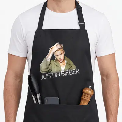 Justin jacket aprons - Justin Bieber photo in olive jacket with white text banner across bottom — shown on a black cotton canvas apron against a white background
