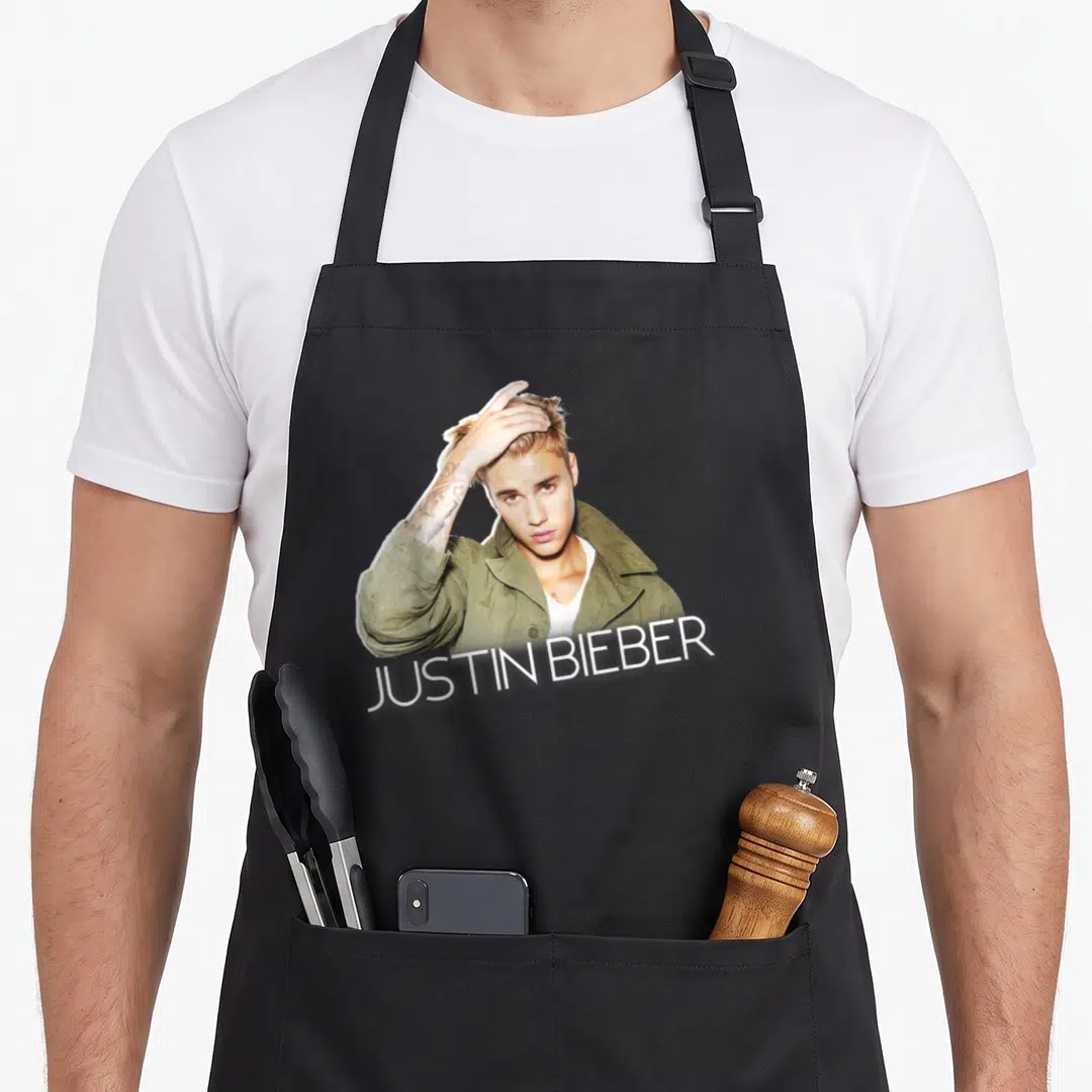 Justin jacket aprons - Justin Bieber photo in olive jacket with white text banner across bottom — shown on a black cotton canvas apron against a white background
