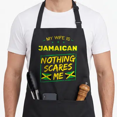 Jamaican wife apron - Stacked yellow and green text reading My Wife Is Jamaican Nothing Scares Me with two flags — shown on a black cotton canvas apron against a white background