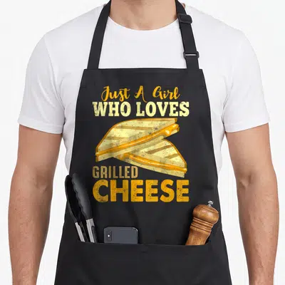 Funny grilled cheese sandwich apron - Orange and yellow text Just A Girl Who Loves Grilled Cheese above two sandwich — shown on a black cotton canvas apron against a white background
