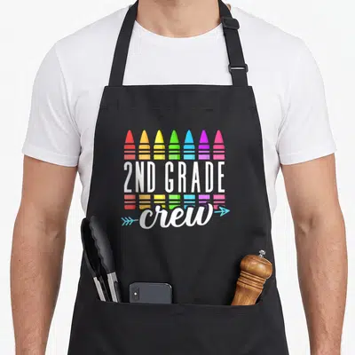 Crew school 2nd apron - Seven crayons in rainbow colors with 2ND GRADE crew text and light blue arrows — shown on a black cotton canvas apron against a white background