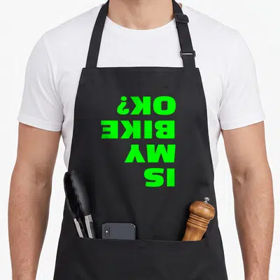 Bike joke apron - Green text reading IS MY BIKE OK? printed upside down on black fabric — shown on a black cotton canvas apron against a white background
