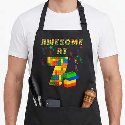 Theme block & apron - Colorful building block letters spelling AWESOME AT 7 with stars confetti and stacked bricks — shown on a black cotton canvas apron against a white background