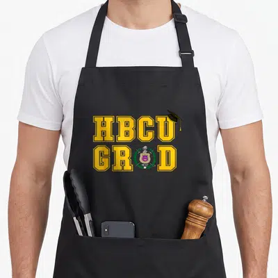 Psi production life apron - HBCU GRAD yellow text with graduation cap and purple shield crest containing swords — shown on a black cotton canvas apron against a white background