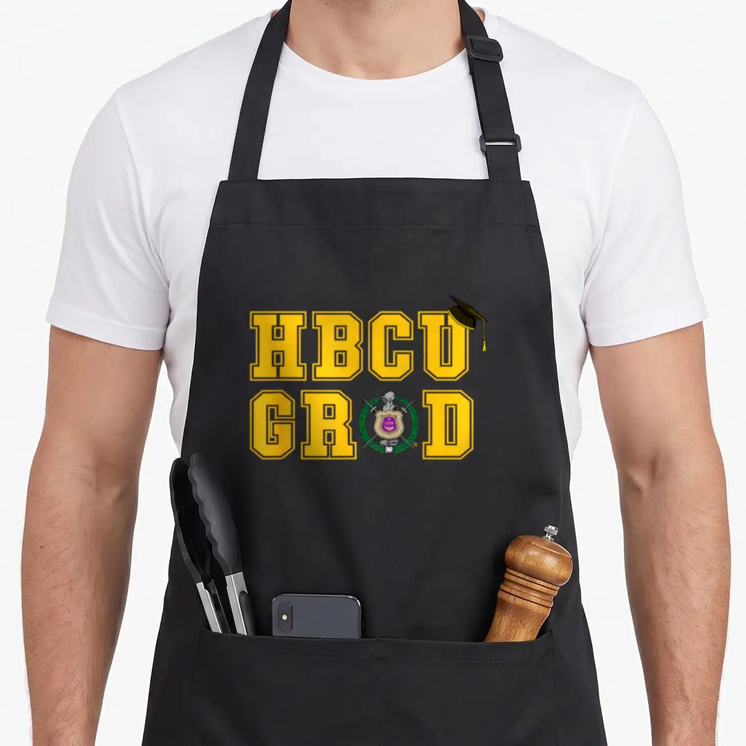 Psi production life apron - HBCU GRAD yellow text with graduation cap and purple shield crest containing swords — shown on a black cotton canvas apron against a white background