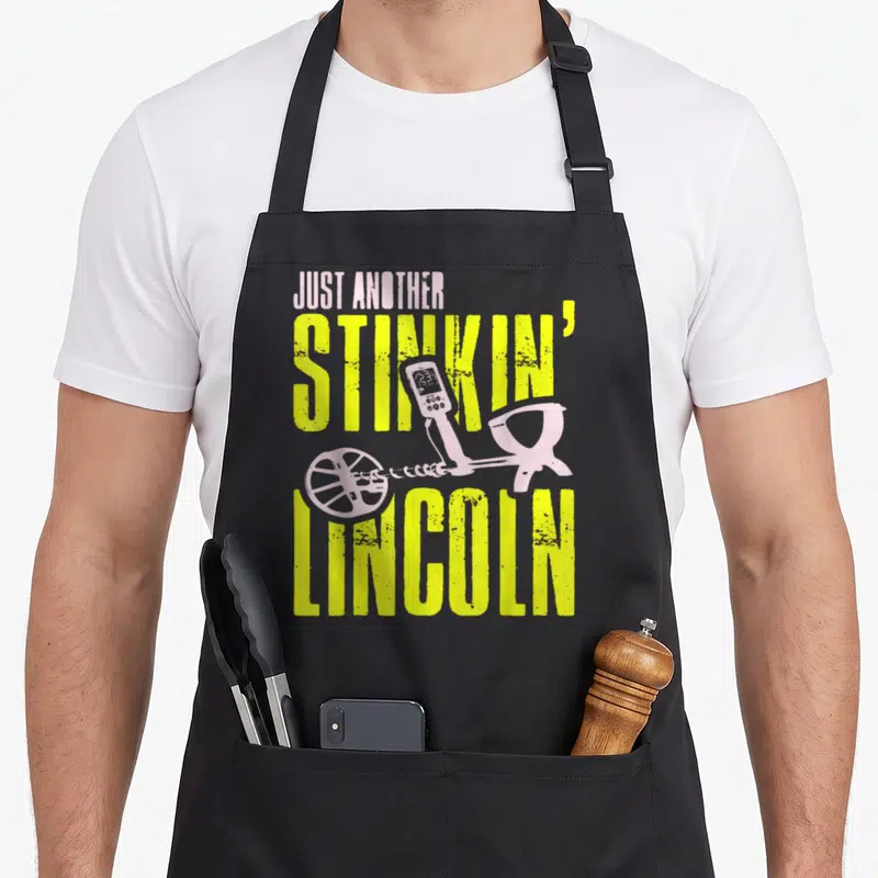 Another metal detecting apron - Yellow distressed text Just Another Stinkin Lincoln with purple metal detector — shown on a black cotton canvas apron against a white background
