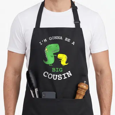 Baby cousin apron - Two cartoon dinosaurs with I'm Gonna Be A Big Cousin text — shown on a black cotton canvas apron against a white background
