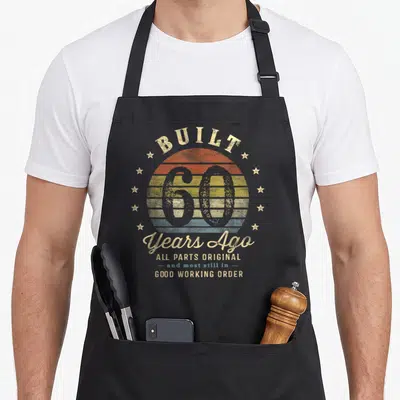 Ago 60th birthday apron - Distressed retro sun with number 60 and text Built 60 Years Ago All Parts Original Good — shown on a black cotton canvas apron against a white background