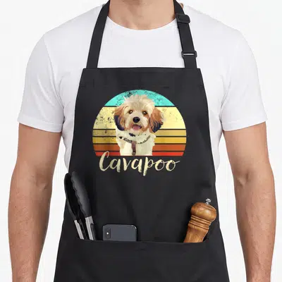 Cavapoo pet dog apron - Retro Cavapoo dog illustration over striped sunset with beige script text — shown on a black cotton canvas apron against a white background