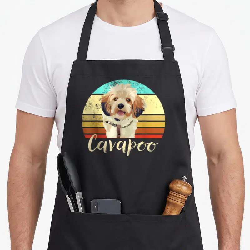 Cavapoo pet dog apron - Retro Cavapoo dog illustration over striped sunset with beige script text — shown on a black cotton canvas apron against a white background
