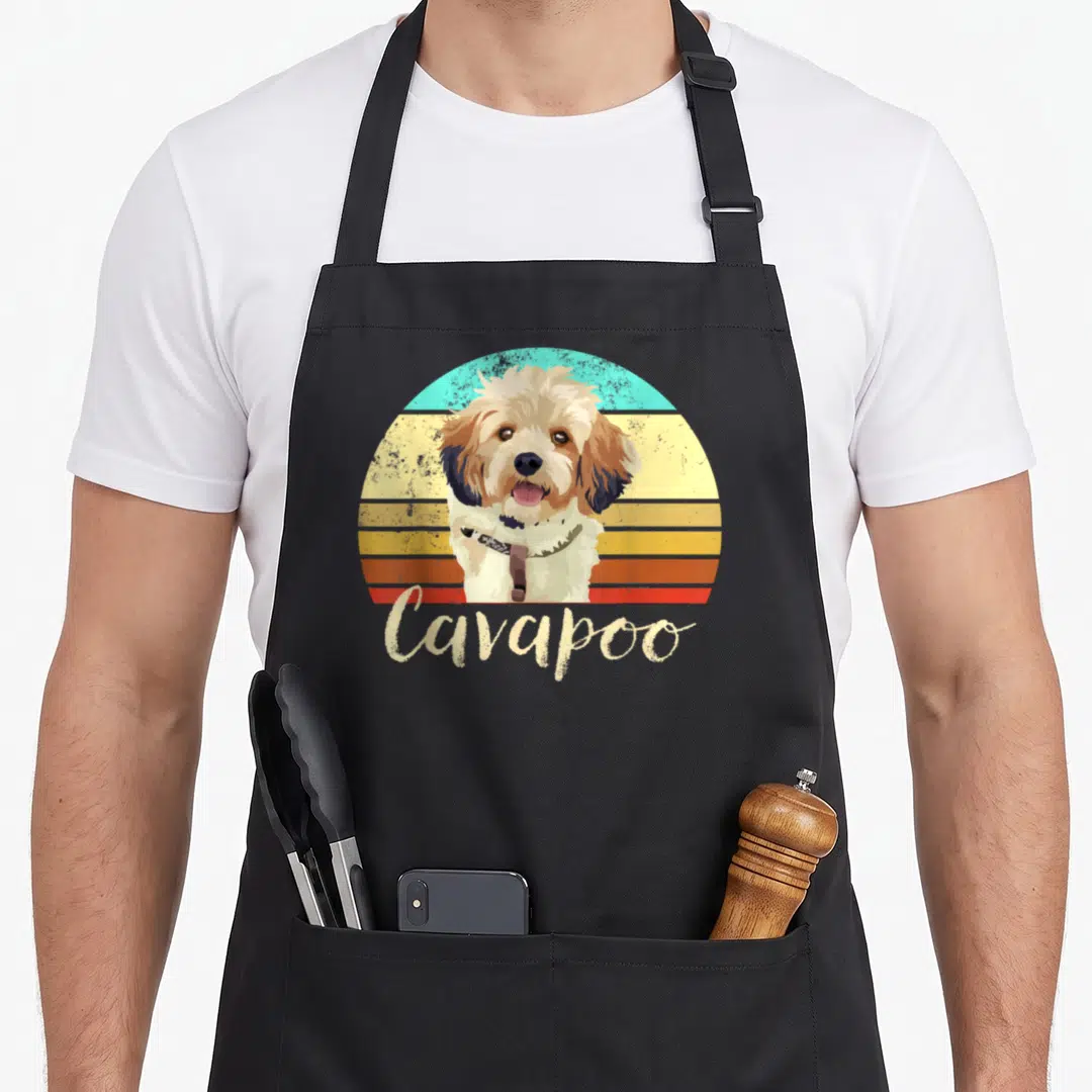 Cavapoo pet dog apron - Retro Cavapoo dog illustration over striped sunset with beige script text — shown on a black cotton canvas apron against a white background