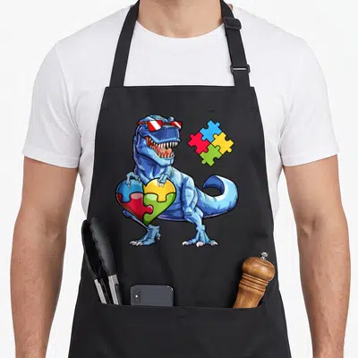 Puzzle autism dinosaur apron - Blue cartoon T-Rex with sunglasses holding a puzzle piece heart made of four colored — shown on a black cotton canvas apron against a white background