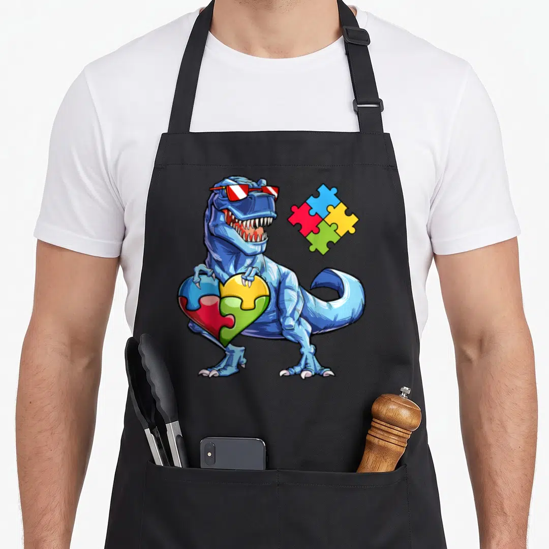 Puzzle autism dinosaur apron - Blue cartoon T-Rex with sunglasses holding a puzzle piece heart made of four colored — shown on a black cotton canvas apron against a white background
