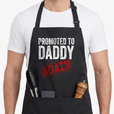 Promoted pregnancy daddy apron - Distressed white text Promoted to Daddy Again with red stamp border on black fabric — shown on a black cotton canvas apron against a white background