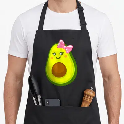 Vintage avocado apron - Cartoon avocado with pink bow, green yellow brown colors, cute face — shown on a black cotton canvas apron against a white background
