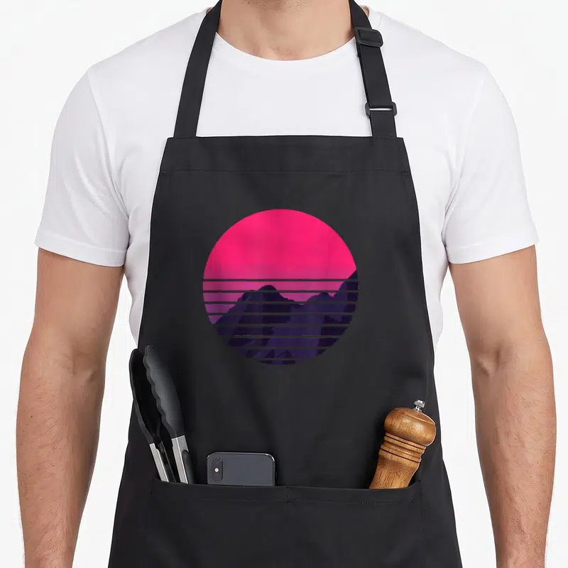 Mountain retro sunset apron - Pink circular sun partially covered by dark purple mountains with horizontal lines — shown on a black cotton canvas apron against a white background