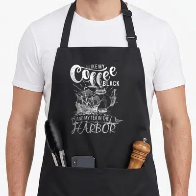 Patriotic coffee aprons - Distressed white text I LIKE MY COFFEE BLACK AND MY TEA IN THE HARBOR with coffee pot — shown on a black cotton canvas apron against a white background