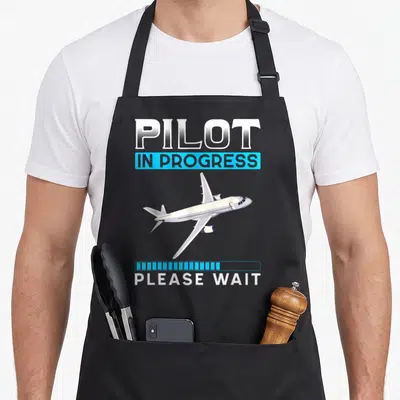 Flying chef and co aprons - White airplane graphic with blue loading bar and Pilot In Progress text — shown on a black cotton canvas apron against a white background