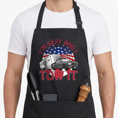 Tow 5th wheel apron - Cartoon truck towing camper under red white blue US flag circle with text I'm Sexy And I Tow It — shown on a black cotton canvas apron against a white background