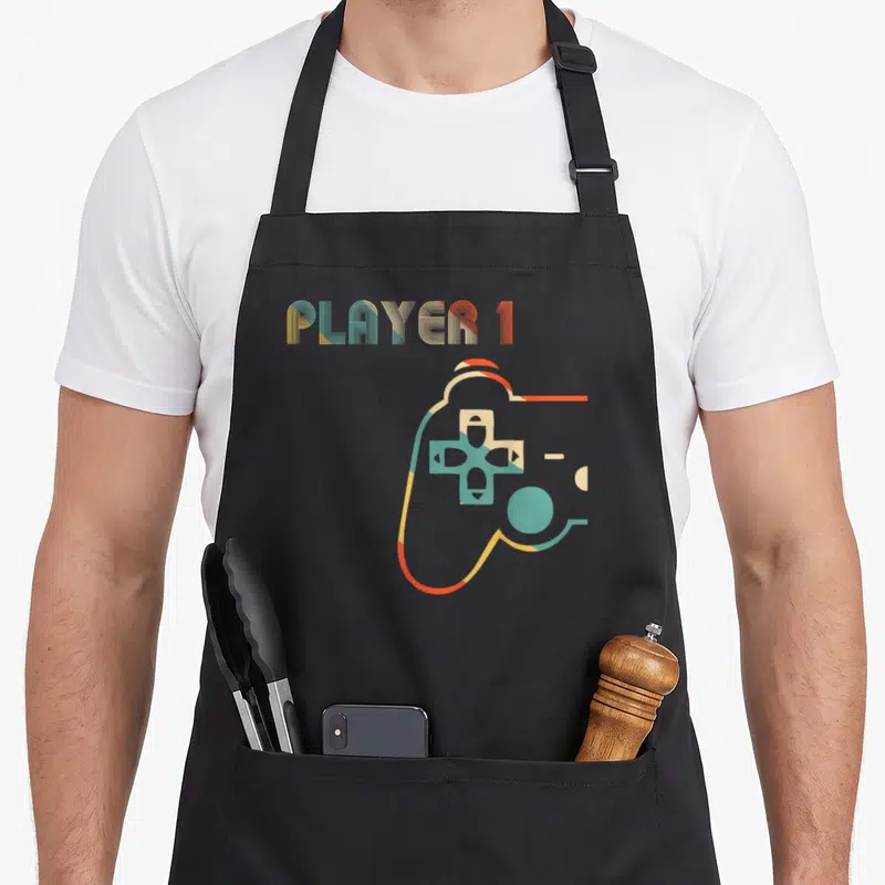 Gamer couple apron - PLAYER 1 text above a red yellow blue green retro game controller outline — shown on a black cotton canvas apron against a white background