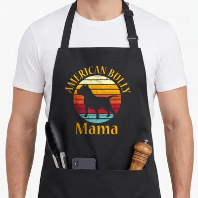 Bully dog apron - Grey American Bully silhouette inside a striped yellow orange red blue sunset circle with Mama text — shown on a black cotton canvas apron against a white background
