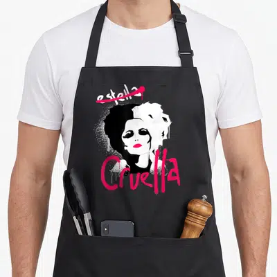 Cruella punk rock apron - Woman with split black and white hair, pink spray paint text estella and Cruella — shown on a black cotton canvas apron against a white background