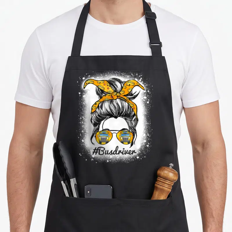 Bus messy aprons - Cartoon messy bun with yellow bus sunglasses and #Busdriver text surrounded by white splatter — shown on a black cotton canvas apron against a white background
