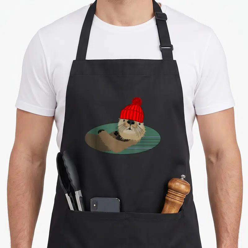 Otter beanie apron - Cartoon brown otter wearing red beanie floating in green oval water — shown on a black cotton canvas apron against a white background