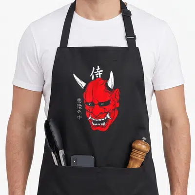Demon face apron - Red oni mask with white horns and Japanese kanji characters samurai and inside demon — shown on a black cotton canvas apron against a white background