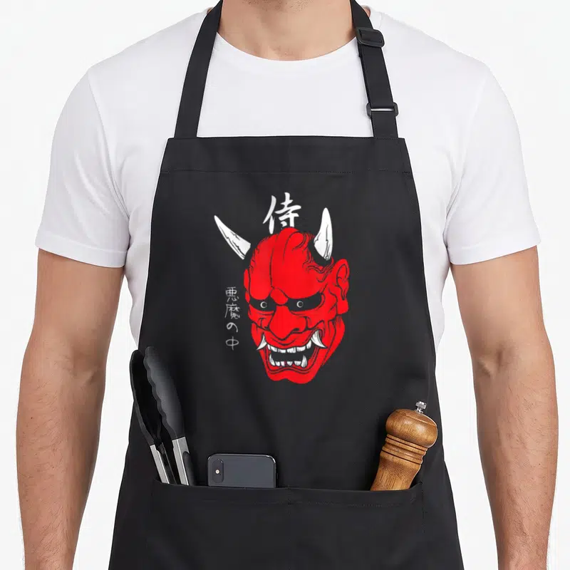 Demon face apron - Red oni mask with white horns and Japanese kanji characters samurai and inside demon — shown on a black cotton canvas apron against a white background