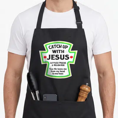 Catch jesus christian apron - Ketchup label graphic with Catch Up With Jesus and two red tomatoes flanking the text — shown on a black cotton canvas apron against a white background