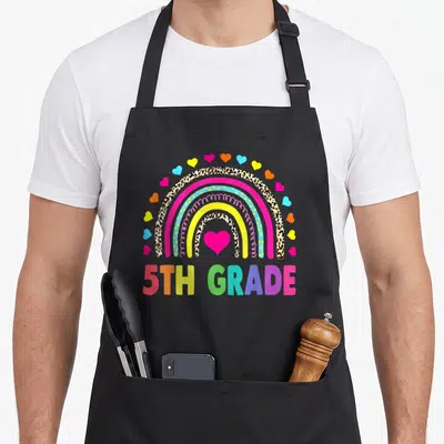 5th rainbow teacher apron - 5TH GRADE text under leopard rainbow with pink heart and scattered colorful hearts — shown on a black cotton canvas apron against a white background