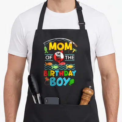 Ally fish apron - Fishing rod arching over MOM THE BIG ONE BIRTHDAY BOY text with colorful cartoon fish and bobber — shown on a black cotton canvas apron against a white background