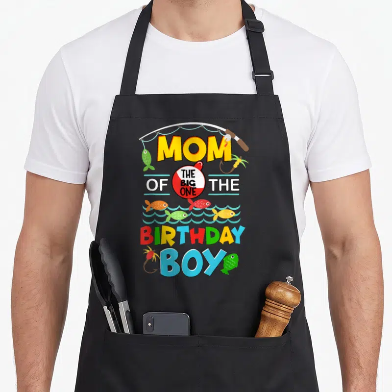 Ally fish apron - Fishing rod arching over MOM THE BIG ONE BIRTHDAY BOY text with colorful cartoon fish and bobber — shown on a black cotton canvas apron against a white background