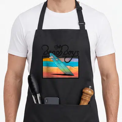Sunset beach apron - Retro surfboard angled over orange and blue sunset stripes with black script text above — shown on a black cotton canvas apron against a white background