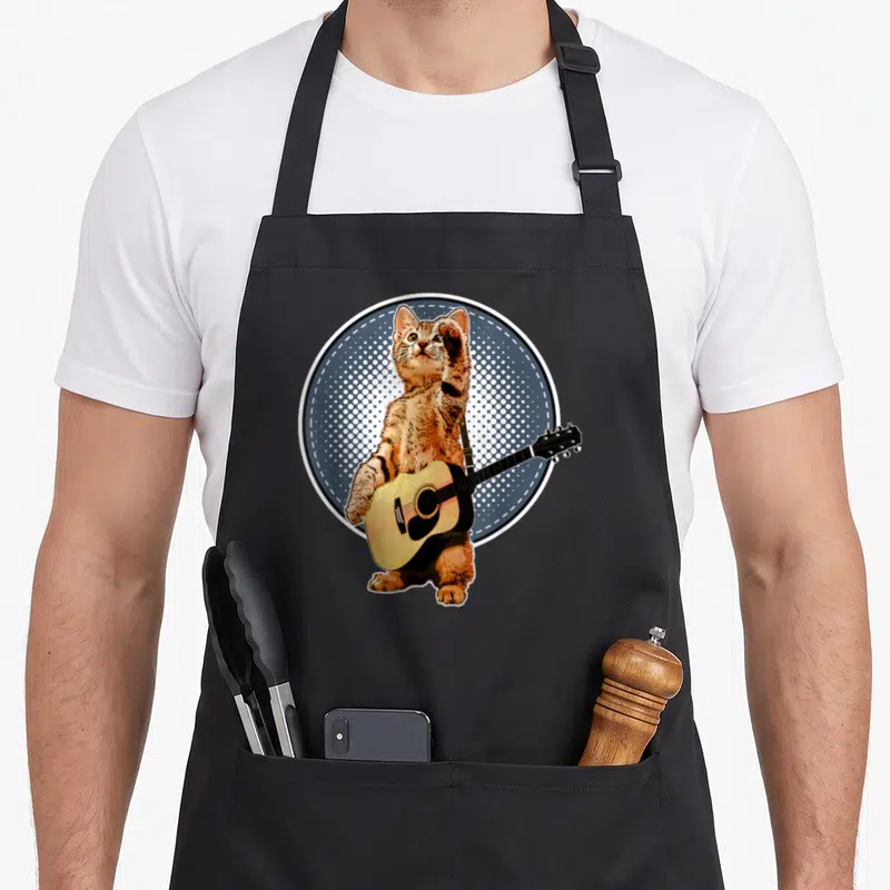 Guitarist cat apron - Orange cat standing holding acoustic guitar in blue halftone circle — shown on a black cotton canvas apron against a white background
