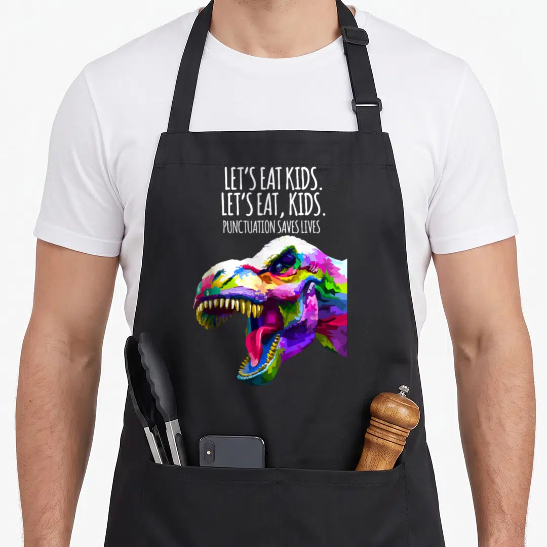 Punctuation save let's eat apron - Colorful pop art T-Rex head below three lines of white text about punctuation — shown on a black cotton canvas apron against a white background