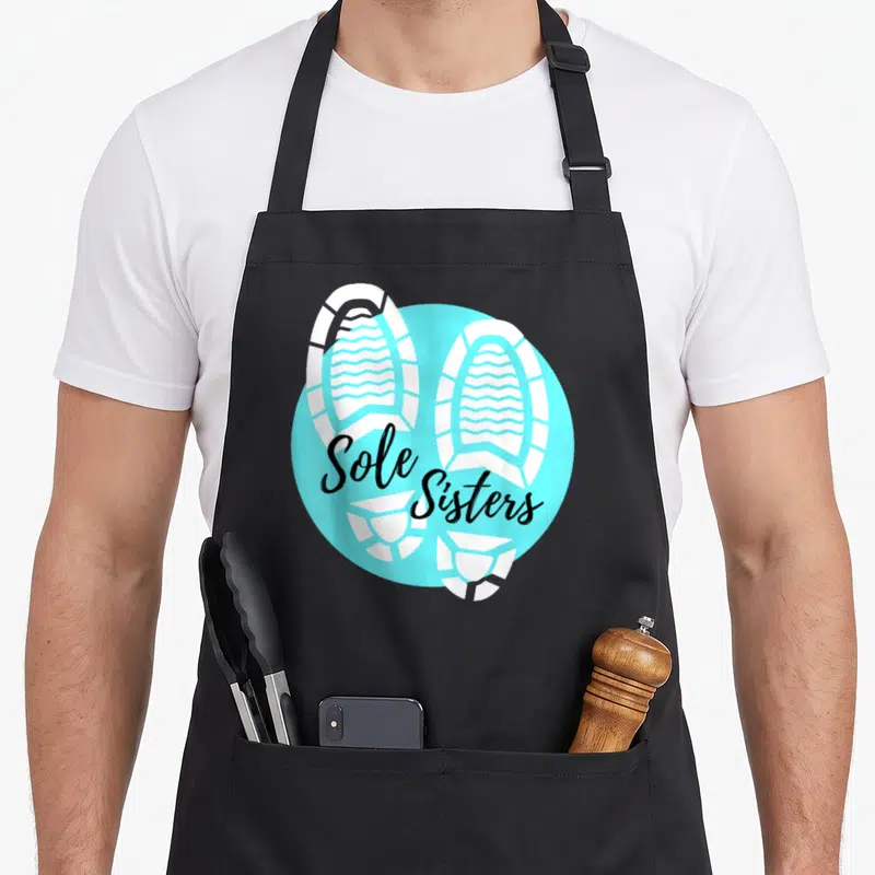 Sole running lover apron - Two white shoe prints on a light blue circle with black Sole Sisters script — shown on a black cotton canvas apron against a white background