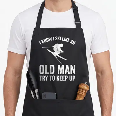 Try ski guy apron - White distressed skier silhouette with text I KNOW I SKI LIKE AN OLD MAN TRY TO KEEP UP — shown on a black cotton canvas apron against a white background
