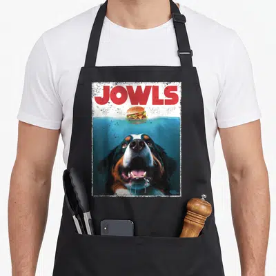 Berner dog apron - Red JOWLS text above a swimming Bernese Mountain Dog reaching for a floating burger — shown on a black cotton canvas apron against a white background