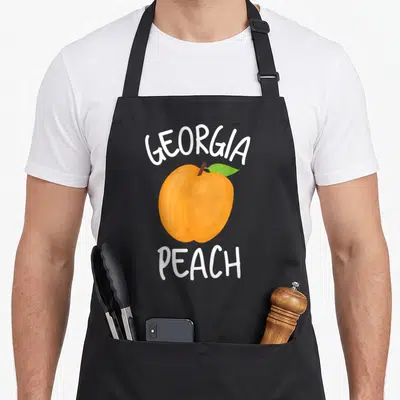 Peach pride apron - Arched GEORGIA text above an orange peach with a green leaf and PEACH below — shown on a black cotton canvas apron against a white background