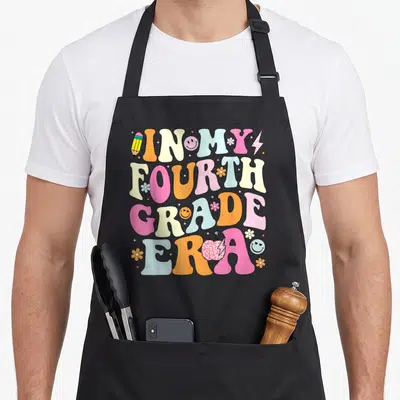 Era 4th teacher apron - In My Fourth Grade Era text in pink, orange, blue, and yellow cartoon letters with retro icons — shown on a black cotton canvas apron against a white background