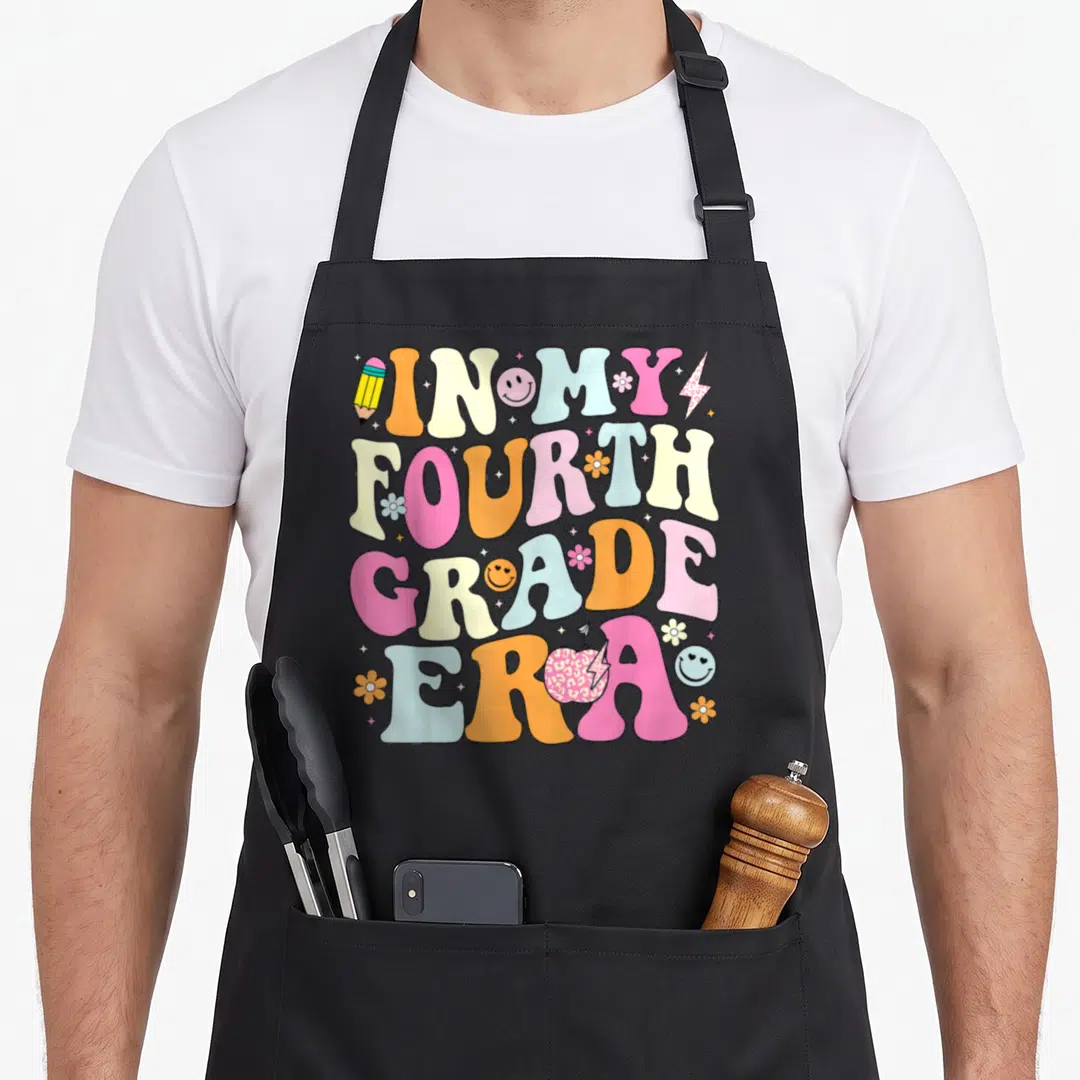 Era 4th teacher apron - In My Fourth Grade Era text in pink, orange, blue, and yellow cartoon letters with retro icons — shown on a black cotton canvas apron against a white background