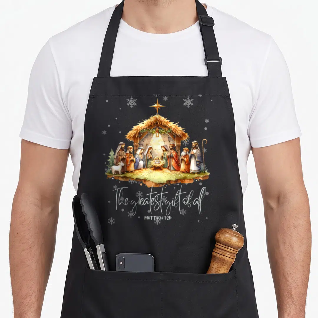 Scene christmas apron - Cartoon nativity with star snowflakes and The greatest gift of all text — shown on a black cotton canvas apron against a white background
